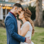 Elopement Wedding A couple dressed in wedding attire stands closely, touching foreheads and smiling. The groom wears a blue suit and maroon shirt, and the bride wears a white lace dress with braided hair. They are outdoors with blurred, glowing string lights in the background, against lush greenery. Elopements Inc